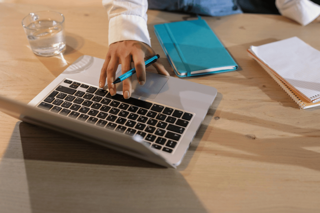 Close-up of hands using a laptop and stylus on a wooden table with a glass of water and notebooks.