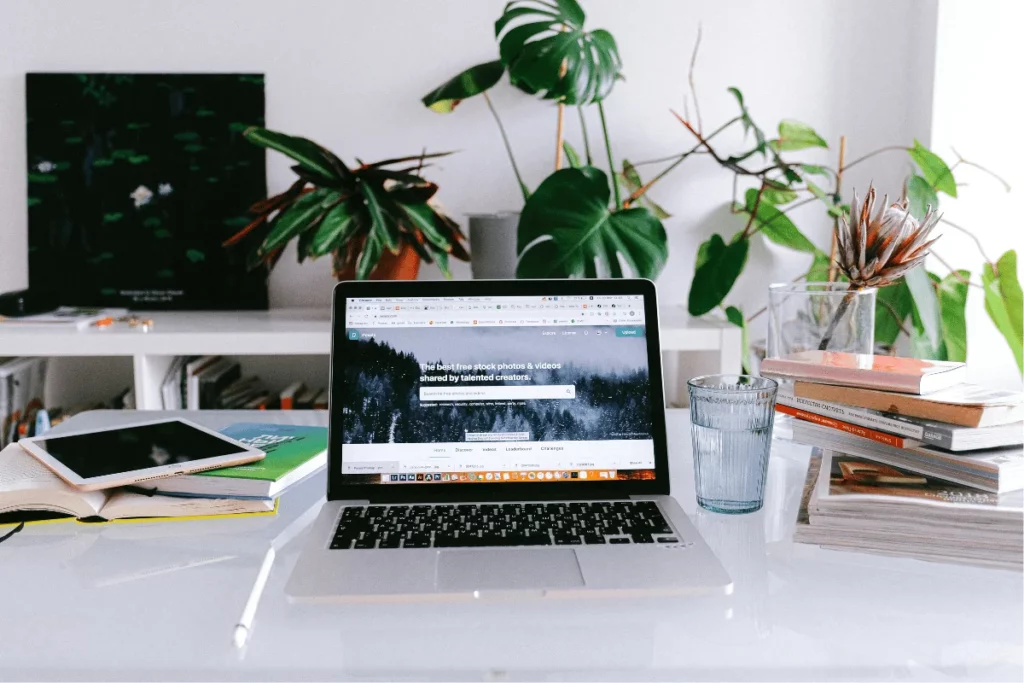 Laptop on a desk with books, a tablet, and a glass of water, surrounded by plants and a bookshelf in the background.