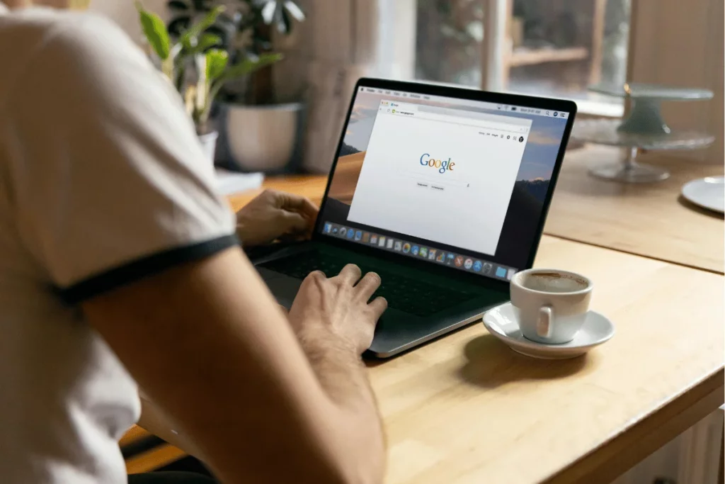 Person using a laptop with Google search page open, while sitting at a wooden table with a cup of coffee.