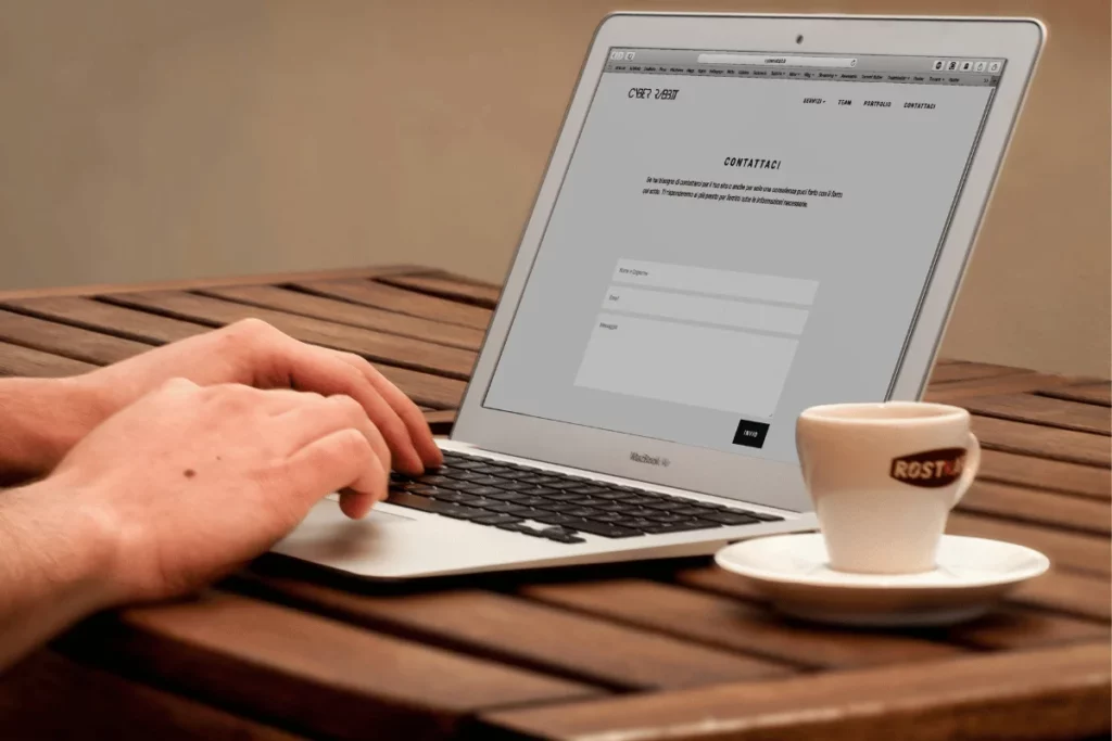 Hands typing on a MacBook Air, filling out a contact form on a website, with a coffee cup on the wooden table.