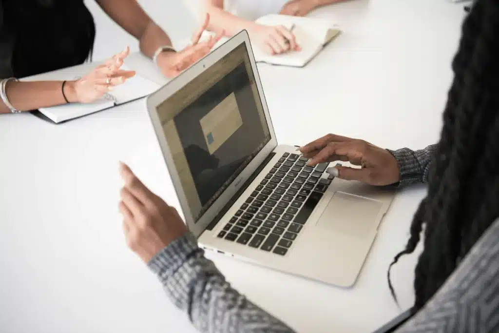 Hands typing on a laptop while discussing project details with team members.