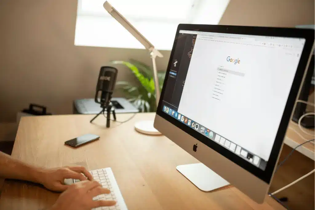 Hands typing on a keyboard with Google search open on an iMac screen.