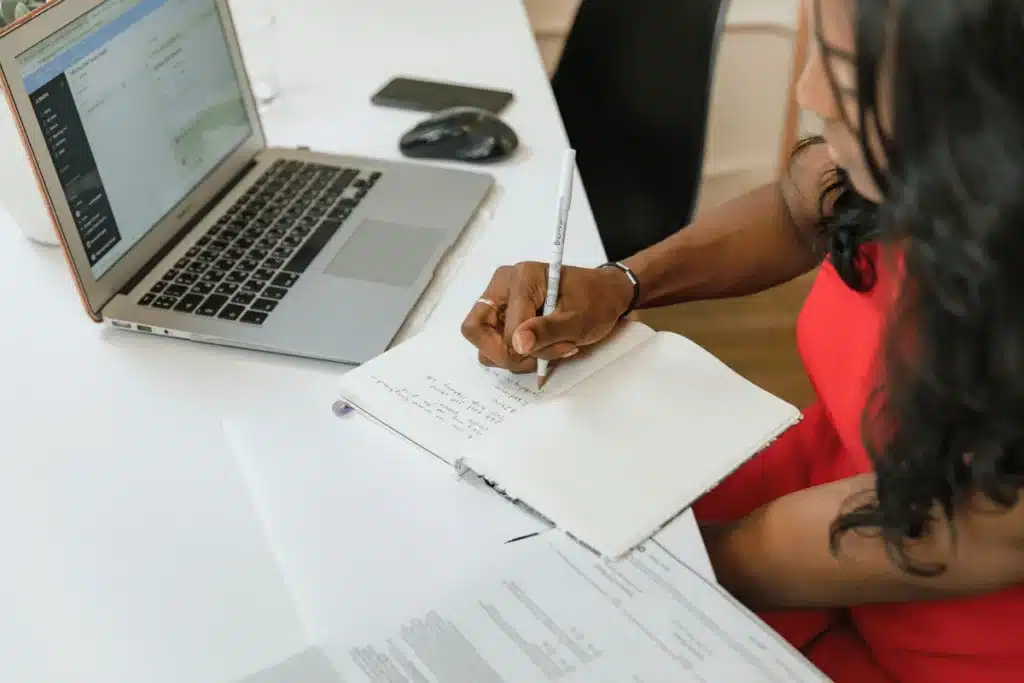 Woman in red dress taking notes beside laptop.