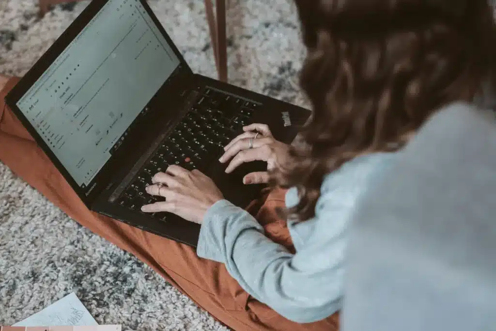 Curly-haired individual typing on a Lenovo laptop on a comfy floor cushion.