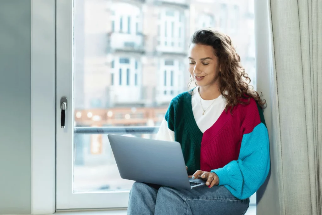 Smiling woman using a laptop while sitting on a windowsill in a bright room.