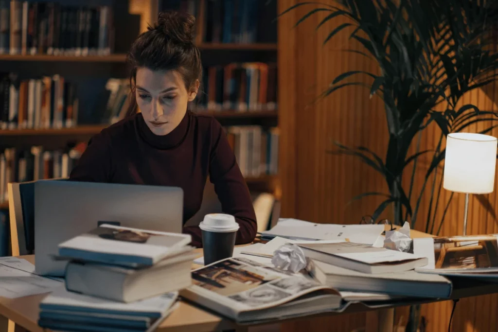 Young woman studying late at night in a library, surrounded by books and papers.