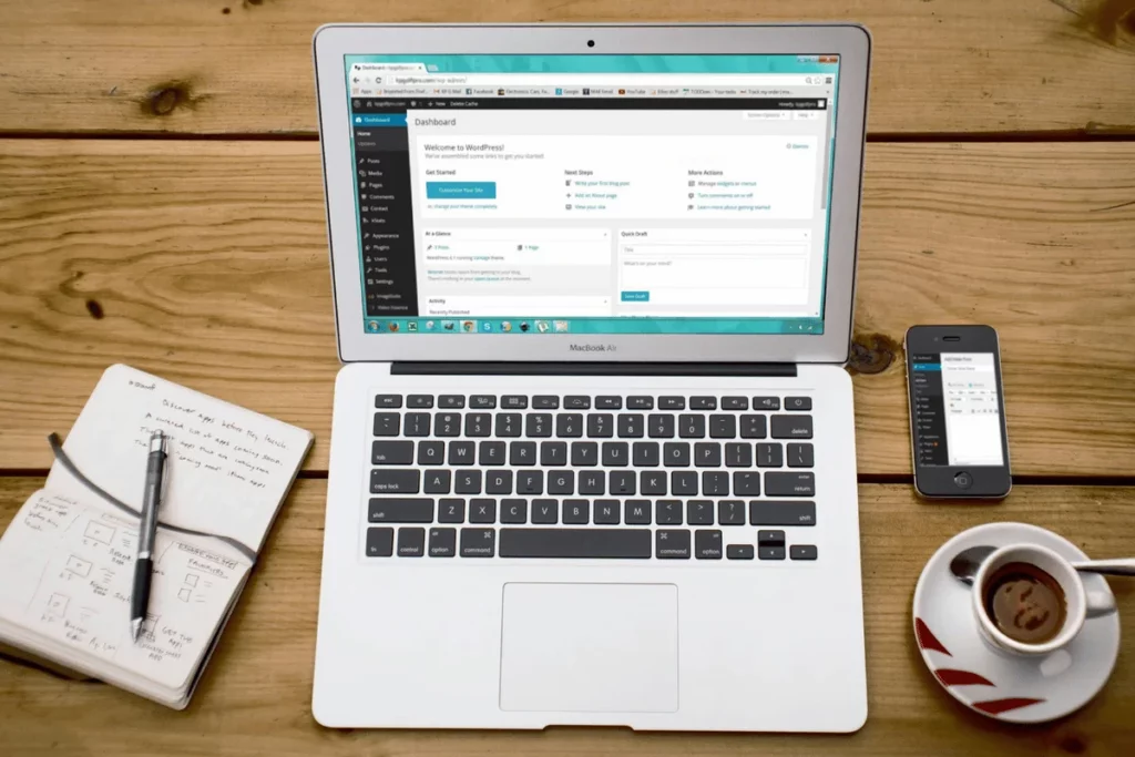 Overhead shot of a wooden desk setup featuring an open laptop with WordPress, a notebook, and a smartphone.