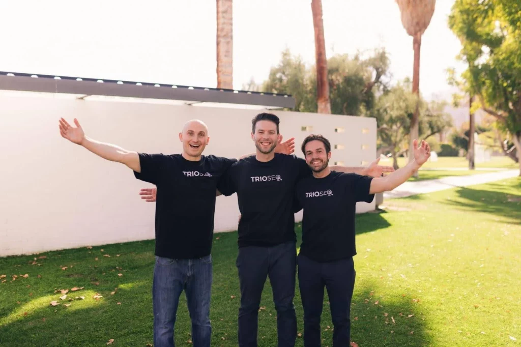 Nathan, Steven and Connor in TRIOSEO shirts stand outdoors with arms outstretched, smiling against a backdrop of grass, palm trees, and a white wall.