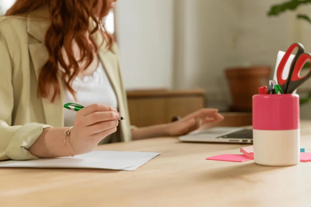 Office scene with a woman preparing to write notes, a laptop and pink sticky notes visible.