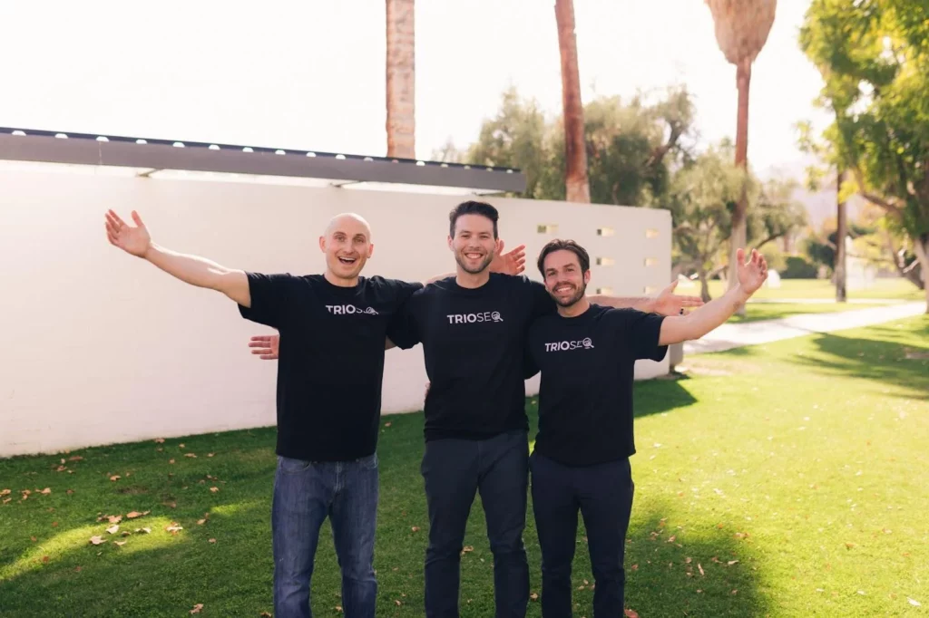 Nathan, Steven and Connor in black t-shirts stand outdoors, arms outstretched, smiling against a backdrop of palm trees and a white wall.