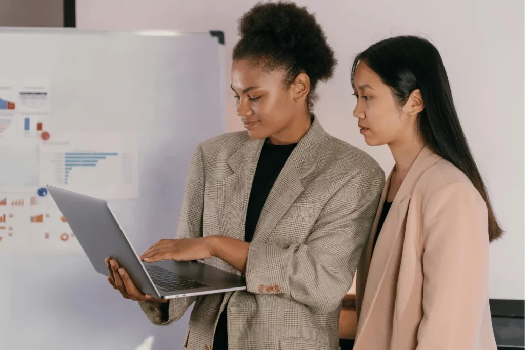 Businesswomen in blazers collaborating on a laptop, with data charts visible on the whiteboard.